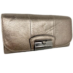May include: A metallic silver Coach wallet with a textured leather finish. The wallet has a rectangular shape with a flap closure secured by a silver-tone clasp. The Coach logo is visible on the clasp.