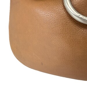 Pu&ograve; includere: Primo piano di una borsa in pelle marrone chiaro con un anello in metallo argentato. La pelle ha una superficie strutturata e una leggera lucentezza. Il design della borsa &egrave; semplice ed elegante.