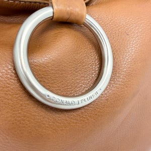 Pu&ograve; includere: Primo piano di una borsa in pelle marrone con una grande maniglia ad anello in metallo argentato. Il marchio "DONALD J PLINER" &egrave; inciso sull'anello. La borsa ha una superficie liscia e strutturata.