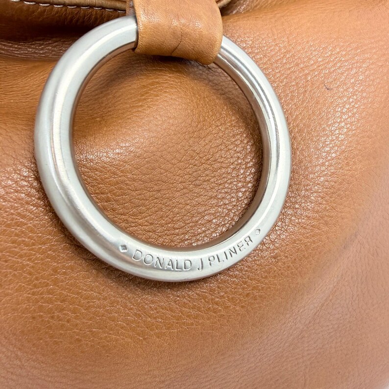 Pu&ograve; includere: Primo piano di una borsa in pelle marrone con una grande maniglia ad anello in metallo argentato. Il marchio "DONALD J PLINER" &egrave; inciso sull'anello. La borsa ha una superficie liscia e strutturata.