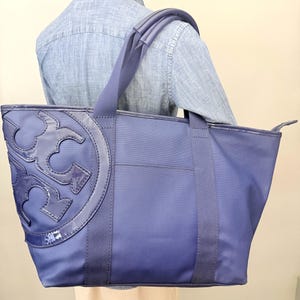 May include: A navy blue tote bag with a large, glossy emblem on the front. The bag has a rectangular shape with a top handle and shoulder strap. The bag is made of a durable material and has a simple, elegant design.