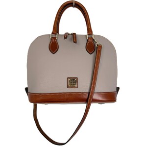 May include: A dome-shaped handbag with a light gray body and brown leather accents. The bag features a top handle, a detachable shoulder strap, and a small gold-colored logo on the front. The base and handle are brown.