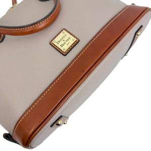 May include: A light gray handbag with brown leather trim and handles. The bag features a stitched design and a small gold-colored plaque that reads "Dooney & Bourke 1975". The bag has a structured shape and a top handle.