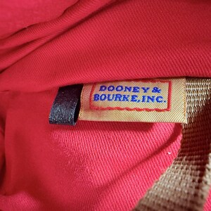 May include: Close-up of a red fabric interior with a tag that reads "DOONEY & BOURKE, INC." in blue text. The tag is rectangular with a red border and a tan background. A black strap is visible.