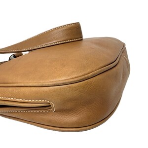 Pu&ograve; includere: Borsa a tracolla in pelle marrone chiaro con un design curvo. La borsa presenta una lunga tracolla e dettagli di cuciture bianche. La pelle ha un aspetto leggermente usurato. La borsa &egrave; su sfondo bianco.