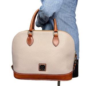 May include: A beige dome-shaped handbag with brown leather accents. The bag features a textured exterior, brown leather handles, and a brown leather base. A small rectangular logo is visible on the front.