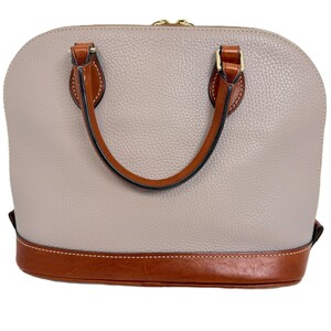 May include: A light gray dome-shaped handbag with brown leather accents. The bag features a textured surface, a brown leather base, and matching handles. Gold-toned hardware secures the handles to the bag.