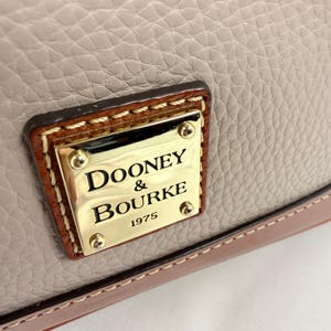 May include: Close-up of a beige leather handbag with a gold metal plaque. The plaque reads "Dooney & Bourke 1975" in black lettering. The bag has brown leather trim and stitching.