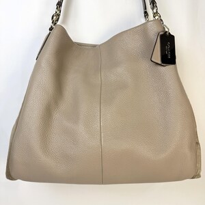 May include: A taupe pebbled leather hobo bag featuring a vertical center seam, silver-toned metal hardware, and a dark brown leather shoulder strap with chain links. A black leather Coach brand tag hangs from the side of the bag.
