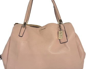 Puede incluir: Un bolso de mano de cuero rosa claro con dos asas superiores y cierre de cremallera. El bolso tiene una superficie texturizada y una pequeña etiqueta dorada con un logotipo. El bolso es de estilo tote.