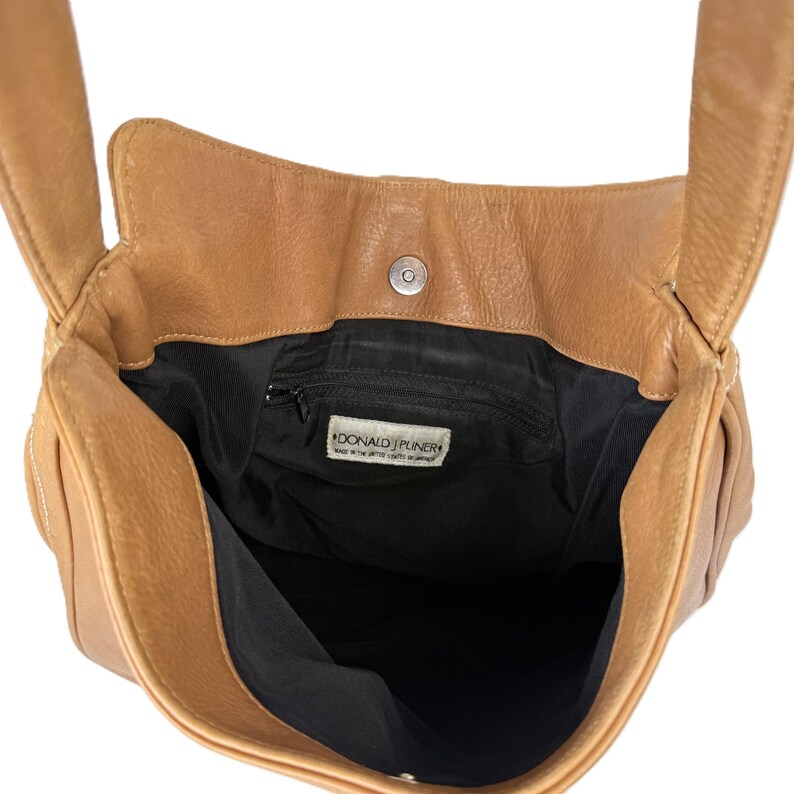 Pu&ograve; includere: Borsa a tracolla in pelle marrone chiaro con interno nero. La borsa ha una chiusura magnetica e una piccola tasca interna con cerniera. Il marchio "DONALD J. PLINER" &egrave; visibile su un'etichetta all'interno della borsa.