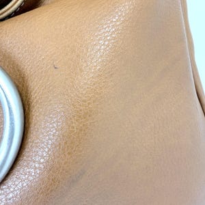 Pu&ograve; includere: Primo piano di una borsa in pelle marrone chiaro. La borsa ha una superficie strutturata e un manico in metallo argentato. La pelle mostra segni di usura, con piccole imperfezioni visibili.
