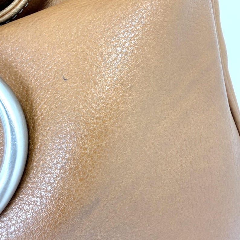 Pu&ograve; includere: Primo piano di una borsa in pelle marrone chiaro. La borsa ha una superficie strutturata e un manico in metallo argentato. La pelle mostra segni di usura, con piccole imperfezioni visibili.