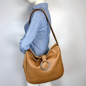 Pu&ograve; includere: Una borsa a tracolla in pelle marrone chiaro con un grande dettaglio ad anello argentato. La borsa ha una lunga tracolla e una cerniera laterale. La borsa &egrave; indossata a spalla.