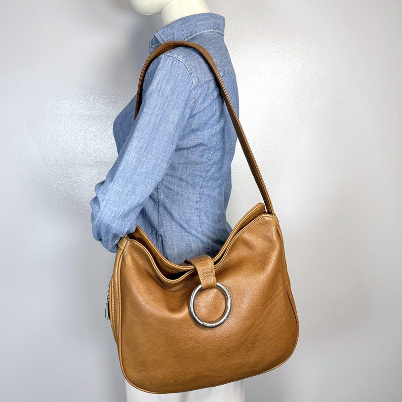Pu&ograve; includere: Una borsa a tracolla in pelle marrone chiaro con un grande dettaglio ad anello argentato. La borsa ha una lunga tracolla e una cerniera laterale. La borsa &egrave; indossata a spalla.
