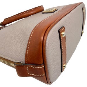May include: A light gray handbag with brown leather trim and a top handle. The bag features gold-colored hardware and white stitching. The bag has a zipper closure.