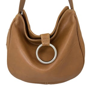 Pu&ograve; includere: Una borsa hobo in pelle marrone chiaro con chiusura ad anello in metallo argentato. La borsa ha una singola tracolla e una forma curva. La pelle sembra morbida e ha una leggera lucentezza. Lo sfondo &egrave; bianco.