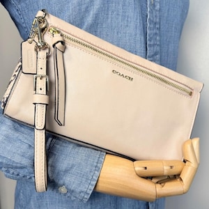 May include: A light beige leather wristlet clutch featuring a gold-tone zipper closure, a matching wrist strap with a buckle, and a gold metal Coach logo on the front. The side panel displays a black and white animal print pattern.