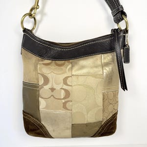 May include: A patchwork shoulder bag with a variety of textures and colors. The bag features a mix of tan, brown, and gold materials, including suede and leather. The bag has a brown leather top and strap, with a gold zipper and brass hardware.