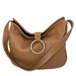 Pu&ograve; includere: Una borsa a tracolla in pelle marrone con chiusura ad anello in metallo argentato. La borsa ha una lunga tracolla e una forma curva. La pelle sembra liscia e ha una leggera lucentezza. La borsa &egrave; vuota e su sfondo bianco.