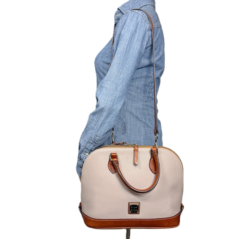 May include: A beige leather satchel with brown leather accents and a brown shoulder strap. The satchel has a rounded shape, a top handle, and a rectangular logo on the front. The bottom of the bag is brown.