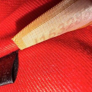 May include: Close-up of a vibrant red fabric with a textured surface. A woven label with a gold and white pattern is partially visible, along with a black label. The image highlights the texture and color of the textile.