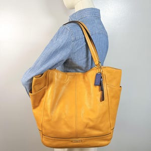 May include: A mustard yellow leather tote bag with two shoulder straps, side pockets, and a blue leather hang tag featuring the Coach brand name. The bag has visible stitching details and a structured base with the Coach logo embossed on the front.
