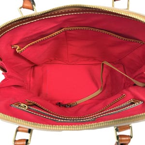 May include: Open view of a beige handbag with brown leather accents and handles. The interior is lined with red fabric and features multiple pockets and a zippered compartment. The bag has a gold zipper closure and a small metal clasp.