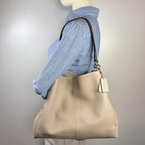 May include: A beige pebbled leather shoulder bag with a slouchy silhouette, featuring a single dark brown shoulder strap attached with silver-toned metal chain links and a rectangular leather luggage tag hanging from the side hardware.