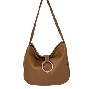 Pu&ograve; includere: Una borsa a tracolla in pelle marrone con una grande chiusura ad anello in metallo argentato. La borsa ha una forma curva e una tracolla lunga e sottile. La pelle sembra liscia e ha una leggera lucentezza. La borsa &egrave; sospesa su sfondo bianco.