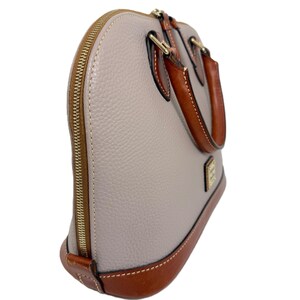 May include: A dome-shaped handbag with a light gray body and brown leather accents. The bag features a gold zipper, handles, and base. The leather handles are stitched to the top of the bag. The bag has a small gold logo on the front.