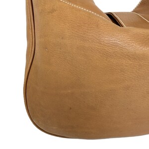 Pu&ograve; includere: Primo piano di una borsa in pelle marrone chiaro. La borsa ha una forma curva con un fondo arrotondato e un cinturino. Le cuciture bianche sono visibili lungo le cuciture. La pelle ha un aspetto leggermente strutturato.