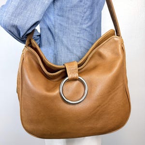 Pu&ograve; includere: Una borsa a tracolla in pelle marrone chiaro con un grande anello argentato. La borsa ha una forma curva e una lunga tracolla. La pelle sembra morbida e usurata. La borsa &egrave; di medie dimensioni, adatta all'uso quotidiano.