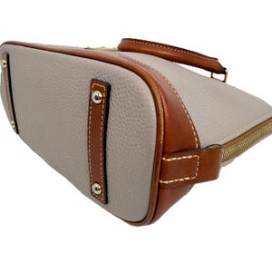 May include: A light gray handbag with brown leather trim and a top handle. The bag features gold-colored hardware, including a zipper and rivets. The leather trim is stitched with white thread, and the bag has a structured shape.