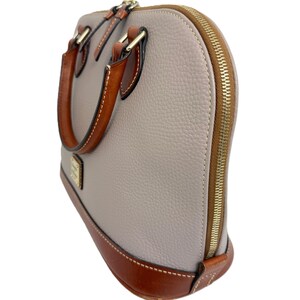 May include: A dome-shaped handbag in a light gray color with brown leather accents. The bag features a gold-tone zipper, handles, and a brown leather base. The handles are stitched with white thread. The bag has a small gold-tone logo on the front.