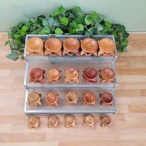 May include: A tiered display rack holds multiple small, wooden sphere stands. The stands are various shades of brown, from light tan to dark reddish-brown. The stands have a round top and a three-legged base. The background includes greenery and a light-colored wooden surface.
