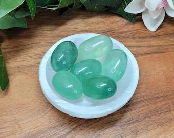 May include: A collection of six smooth, polished, oval-shaped green aventurine stones resting inside a small, round, white selenite bowl. The stones exhibit varying shades of translucent light to deep forest green with a glossy, reflective surface.