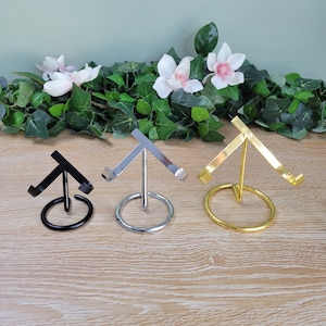 May include: Three metal display stands in black, silver, and gold. Each stand has a circular base and a triangular top, designed to hold items. The stands are arranged on a wooden surface, with greenery and white flowers in the background.