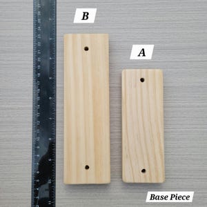 Puede incluir: Dos piezas de base rectangulares de madera clara etiquetadas como A y B, colocadas junto a una regla negra. La pieza B mide aproximadamente 26,7 cm de largo y la pieza A aproximadamente 21,6 cm. Ambas piezas tienen dos orificios circulares.
