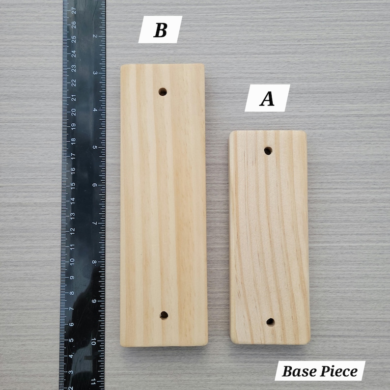 Puede incluir: Dos piezas de base rectangulares de madera clara etiquetadas como A y B, colocadas junto a una regla negra. La pieza B mide aproximadamente 26,7 cm de largo y la pieza A aproximadamente 21,6 cm. Ambas piezas tienen dos orificios circulares.