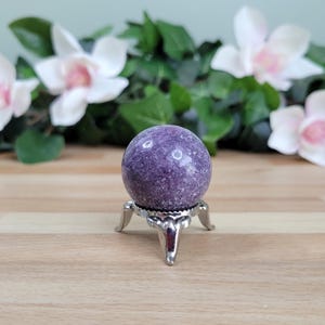 May include: A purple lepidolite sphere on a silver stand. The sphere is smooth and has a speckled appearance.