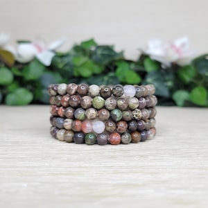 May include: A stack of beaded bracelets in various earth tones. The bracelets feature a mix of brown, green, red, and white beads. The beads are round and appear to be made of stone or a similar material. The bracelets are stacked on a light-colored wooden surface.