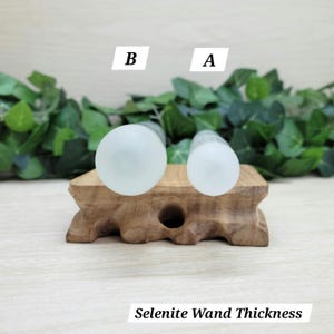 Puede incluir: Dos varitas cil&iacute;ndricas de selenita colocadas verticalmente sobre un soporte de madera, etiquetadas como A y B para mostrar el grosor. Las varitas son de color blanco transl&uacute;cido con una textura lisa. El texto Selenite Wand Thickness es visible.