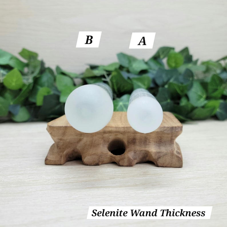 Puede incluir: Dos varitas cil&iacute;ndricas de selenita colocadas verticalmente sobre un soporte de madera, etiquetadas como A y B para mostrar el grosor. Las varitas son de color blanco transl&uacute;cido con una textura lisa. El texto Selenite Wand Thickness es visible.