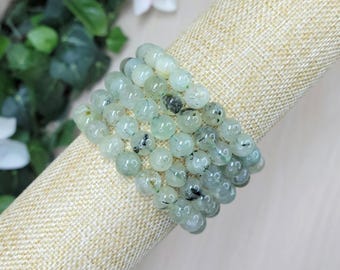 May include: A stretch bracelet composed of multiple rows of round, pale green beads. Each bead features a smooth, polished finish with visible dark, needle-like inclusions scattered throughout the translucent, light green stone material.