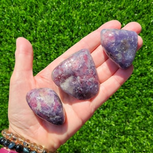 May include: Three tumbled stones in shades of purple and pink are held in a hand against a backdrop of green grass. The stones have a smooth, polished appearance, with variations in color and texture. The stones are likely used for crystal healing or decorative purposes.