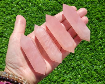 May include: Four double-terminated rose quartz crystal points, each featuring a smooth, translucent, pale pink surface with a hexagonal shape, held in a palm to display their elongated, faceted structure and light-reflecting properties.