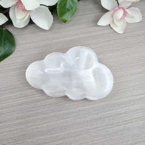 May include: A white selenite crystal dish carved into a cloud shape with a smooth, polished surface and subtle translucent banding. The object features a rounded, organic silhouette with multiple curved edges, reflecting light across its surface.