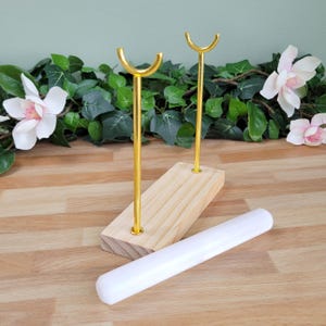 Puede incluir: Un soporte de exhibici&oacute;n con una base rectangular de madera clara y dos varillas met&aacute;licas verticales de tono dorado rematadas con soportes en forma de U, junto a una varilla cil&iacute;ndrica blanca lisa sobre la superficie.