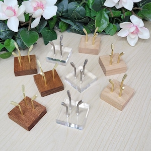 May include: A collection of small display stands. Some stands are made of clear acrylic, others of wood in various shades of brown. Each stand features metal display arms with a curved design, perfect for showcasing small items.